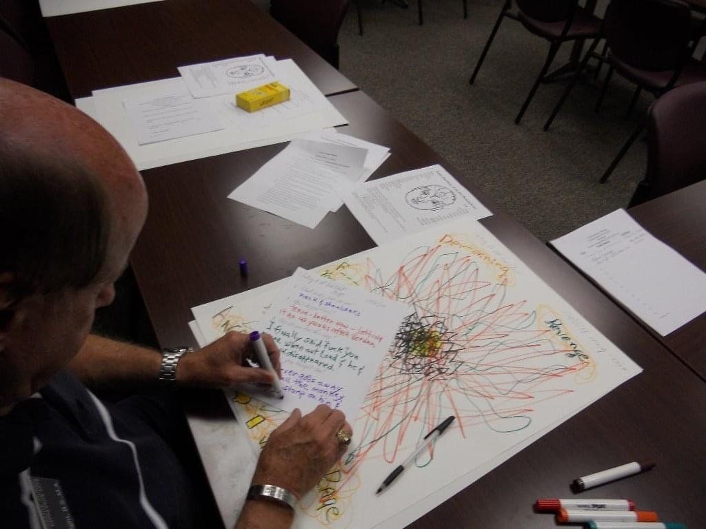 Man writing on a large drawing of a flower with papers on a table, markers, and a yellow block.