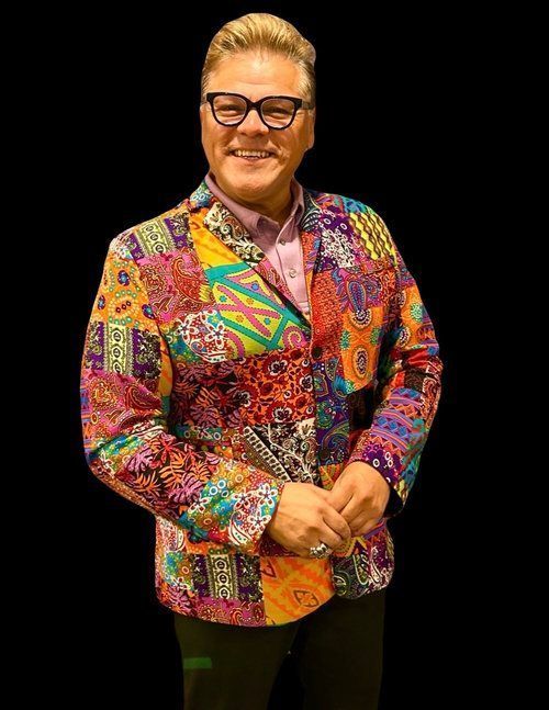 Man in a colorful patchwork blazer smiles, wearing glasses, hands clasped, black background.
