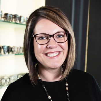 A woman wearing glasses and a necklace is smiling in front of a display of glasses.