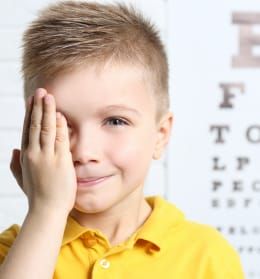 A young boy in a yellow shirt is covering his eye with his hand.