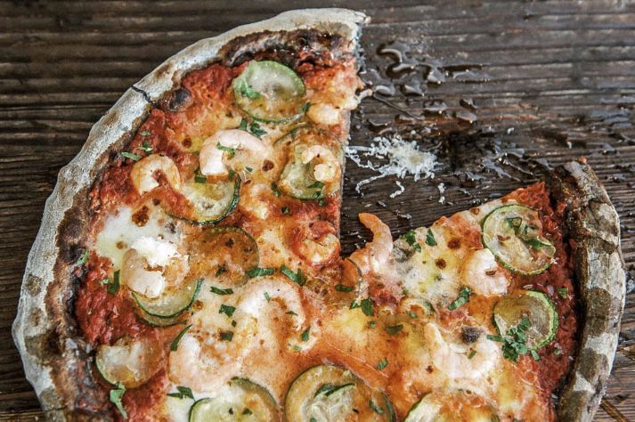 A pizza with shrimp and zucchini is on a wooden table with a slice taken out of it.