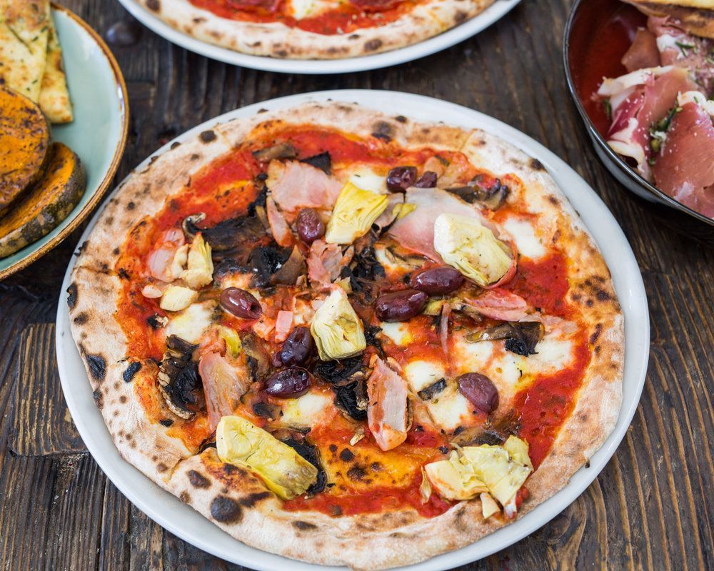 A pizza with artichokes , olives and ham on a white plate on a wooden table.