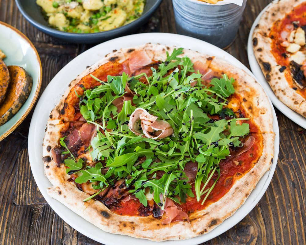 A pizza with a lot of greens on it is on a white plate on a wooden table.