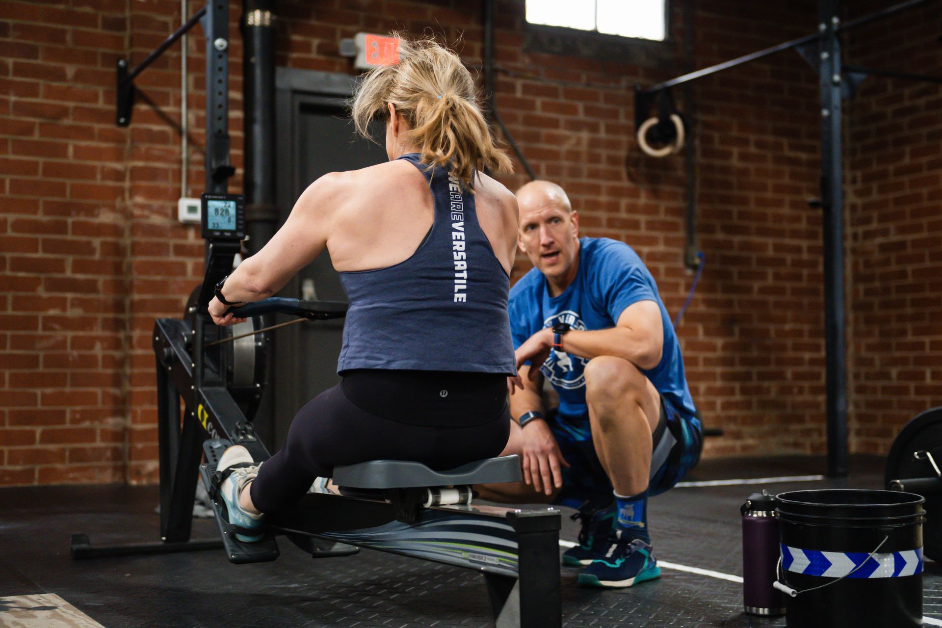 A man is squatting next to a woman on a rowing machine in a gym.