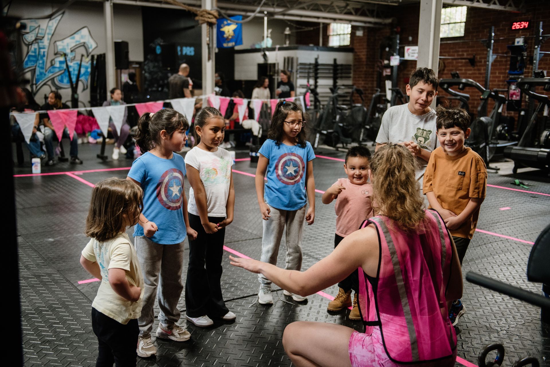 Children in a gym listen to an instructor who points at them, pink lines mark the floor.