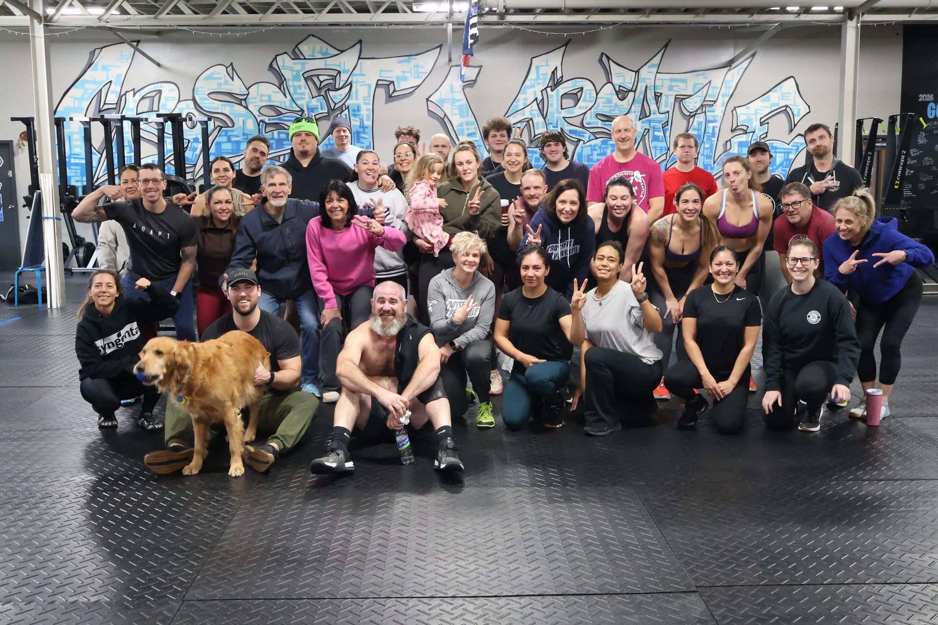 Large group of people pose in gym; some smiling, with dog. Graffiti wall in the background.