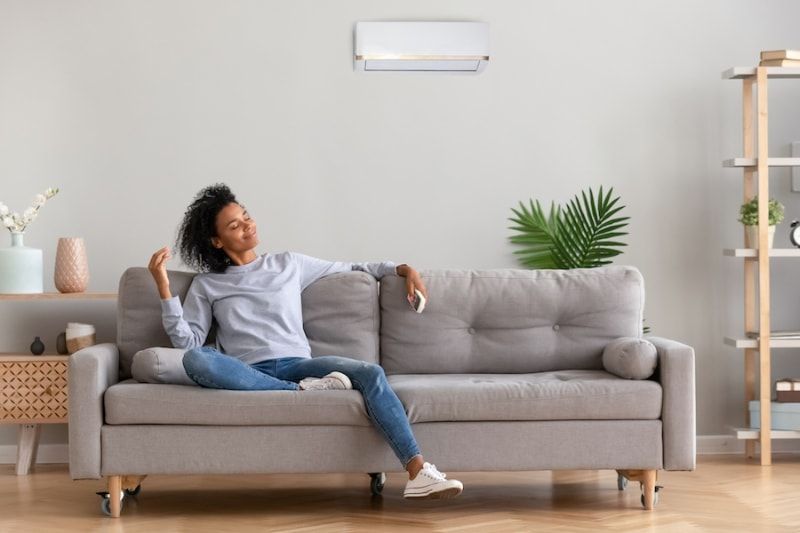 A woman is sitting on a couch in a living room with an air conditioner on the wall.