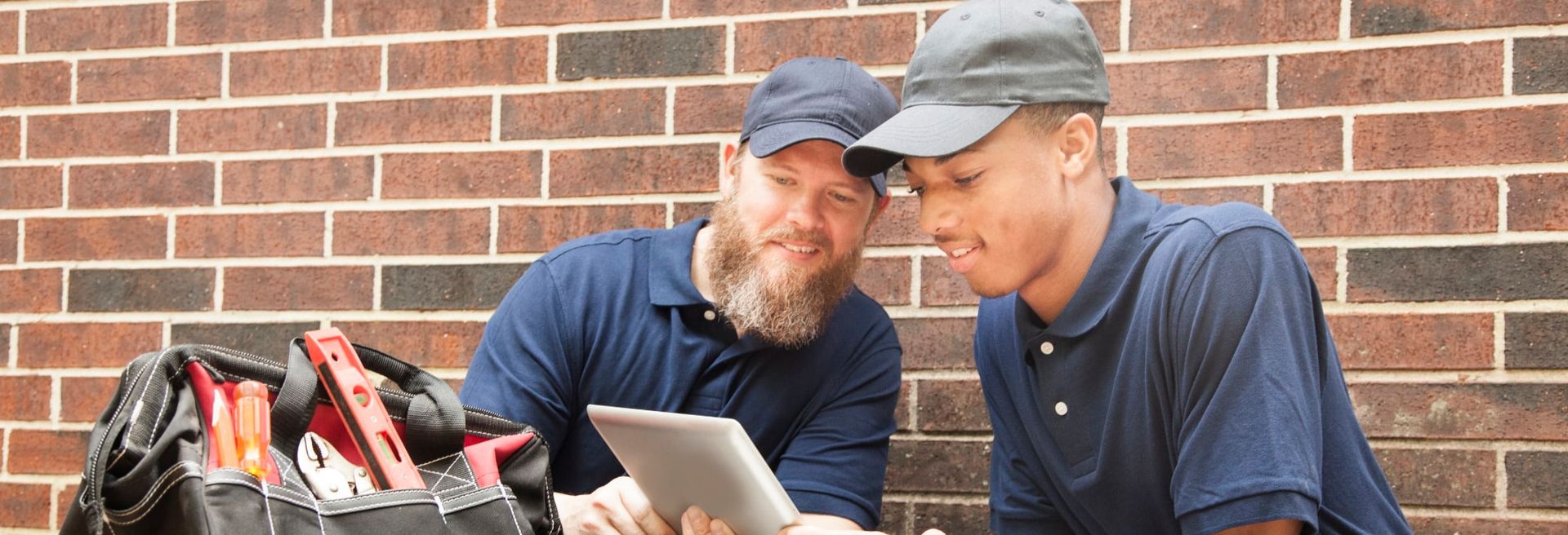 two men are looking at a tablet in front of a brick wall .