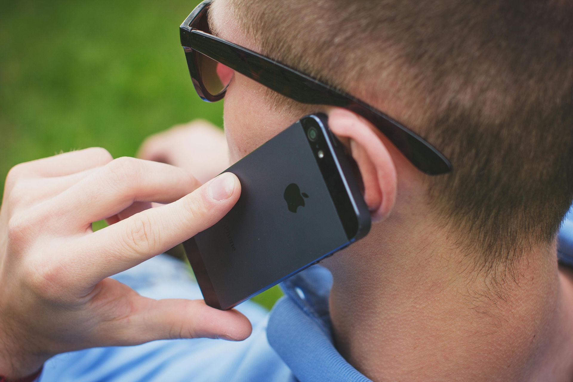 A person wearing sunglasses holding a dark-colored iPhone to their ear outdoors.