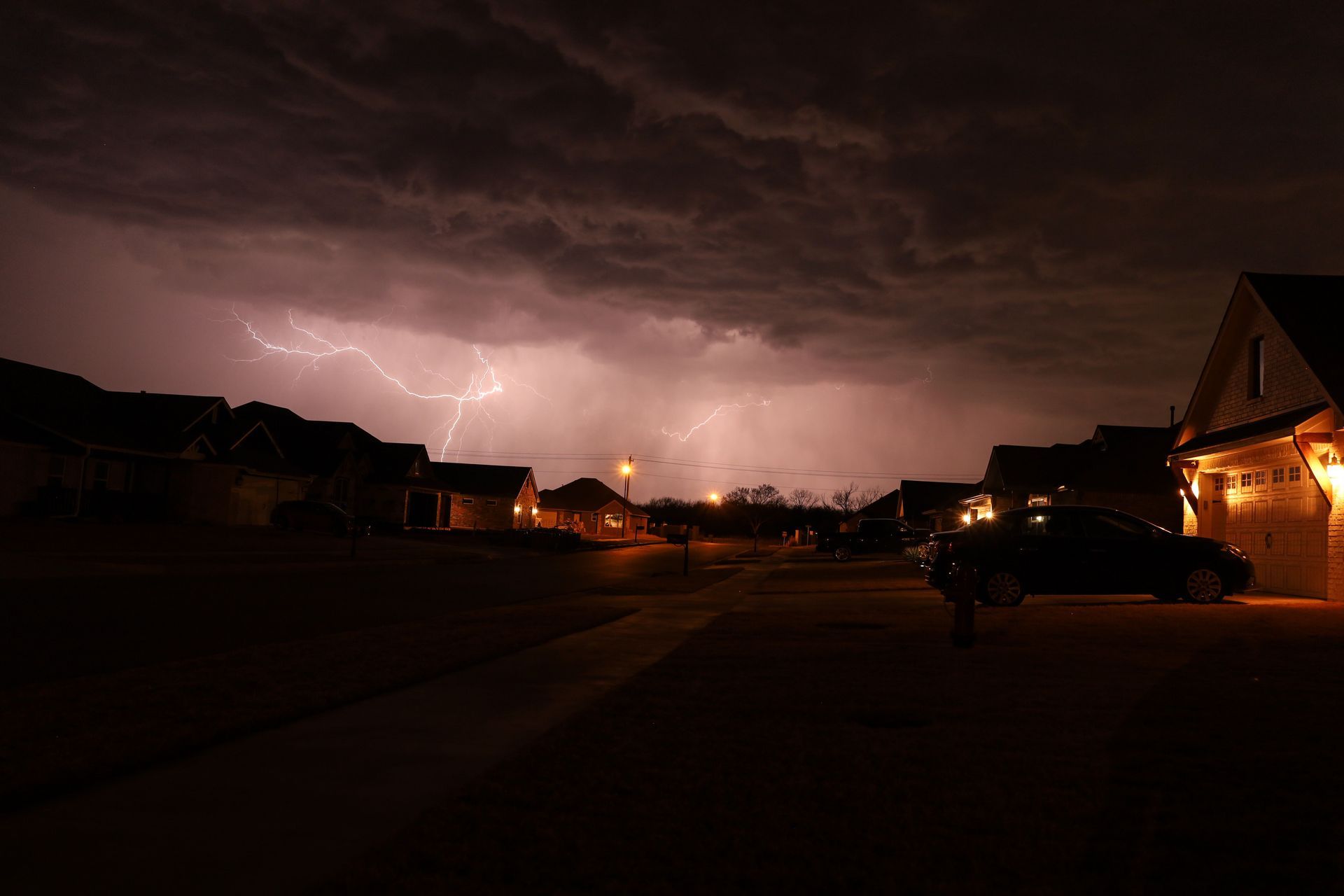 Lightning lights a dark suburban street at night, with glowing house windows under storm clouds.