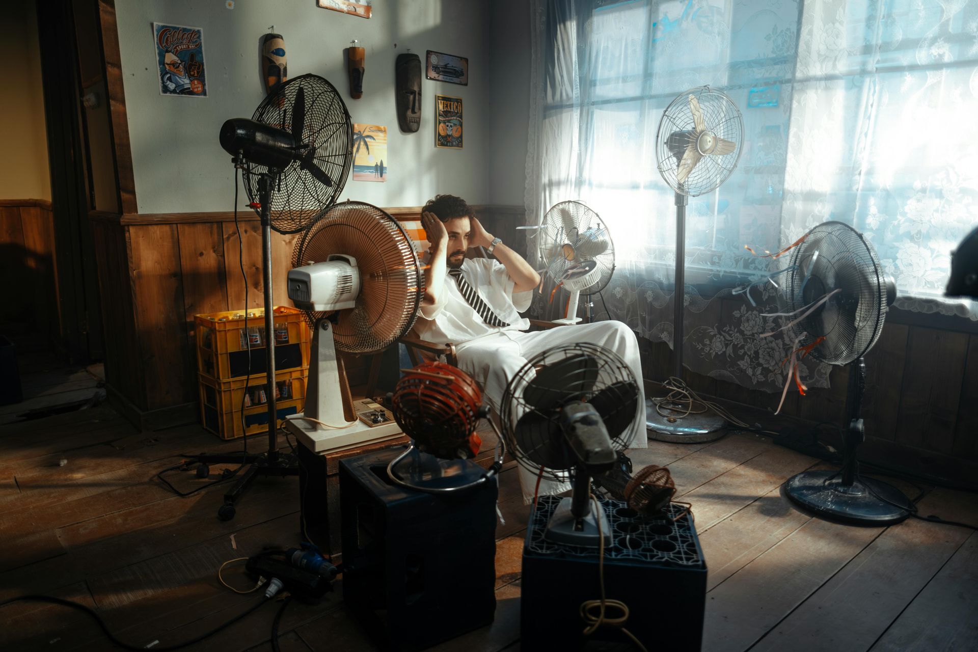 A person sits in a dimly lit room surrounded by multiple electric fans, holding their head with both hands.