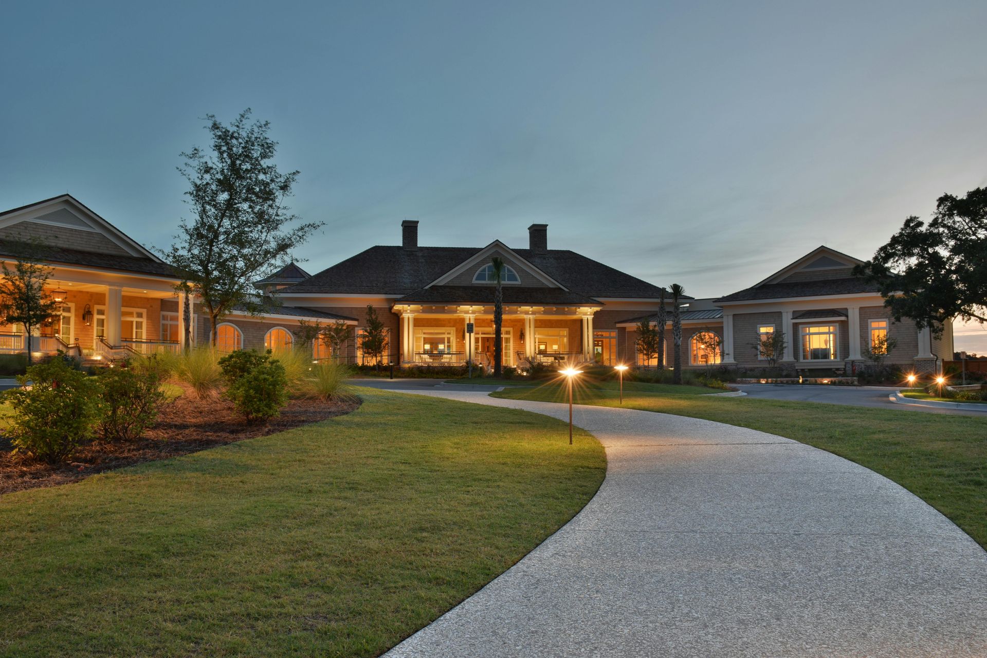 Stone pathway leads to a large, well-lit building with a lawn at dusk.