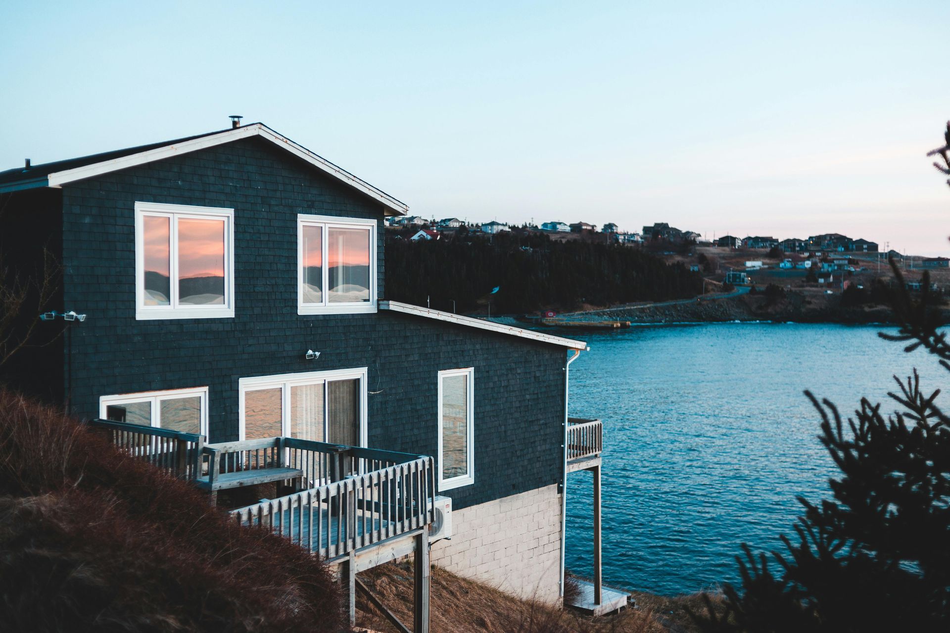 A dark-sided house with a wooden deck overlooking a coastal bay at dusk.