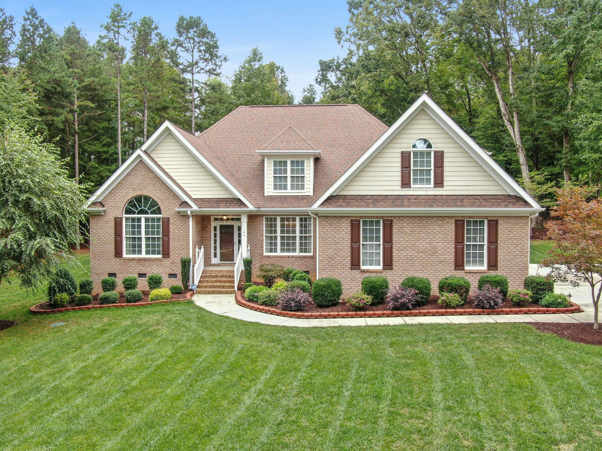 Brick house with tan siding, white trim, and a manicured green lawn surrounded by trees