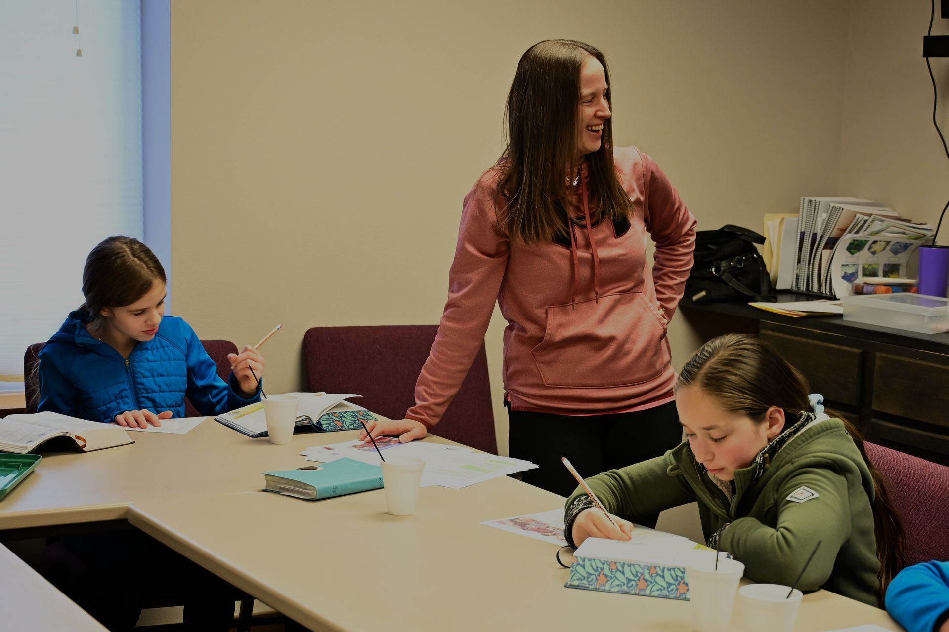 A woman is standing next to two girls sitting at a table in a classroom.