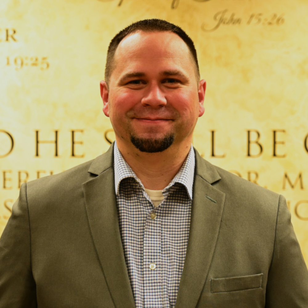 A man in a suit stands in front of a wall that says john 15:26