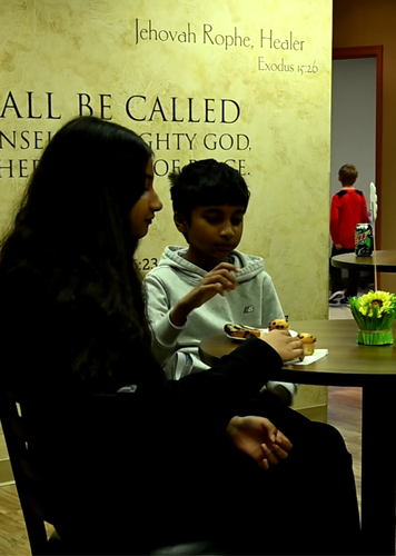 A girl and a boy are sitting at a table in front of a wall that says all be called