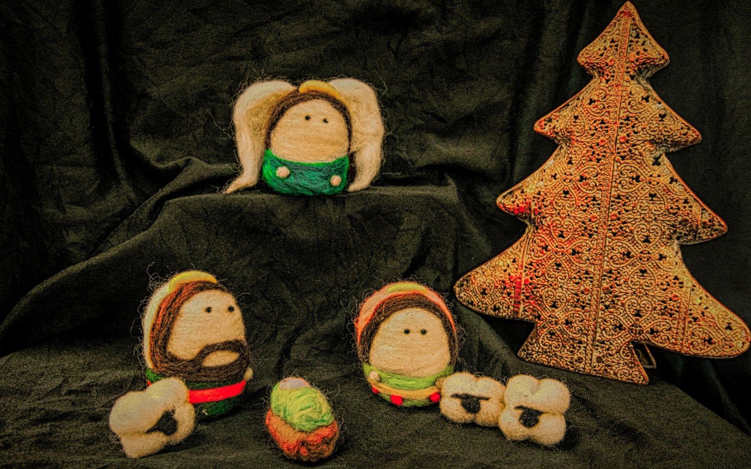 Felt nativity scene: angel, figures, sheep, and Christmas tree against a dark backdrop.
