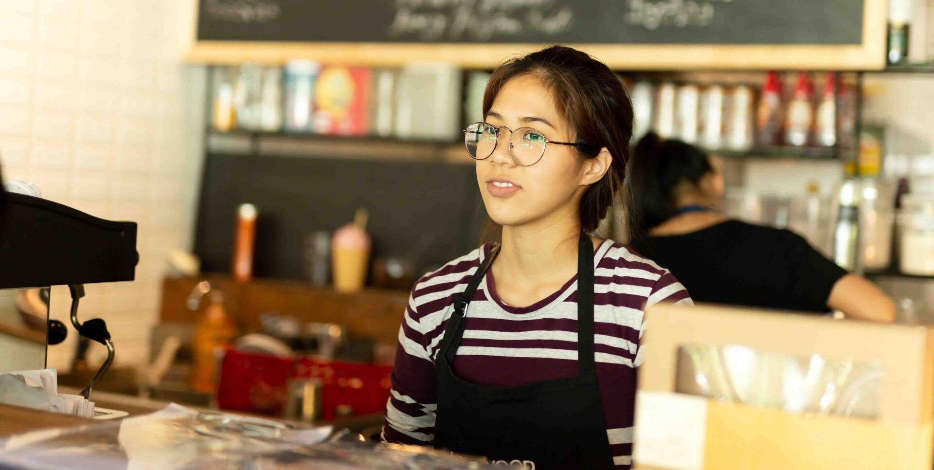 A woman wearing glasses and an apron is sitting at a counter in a restaurant.