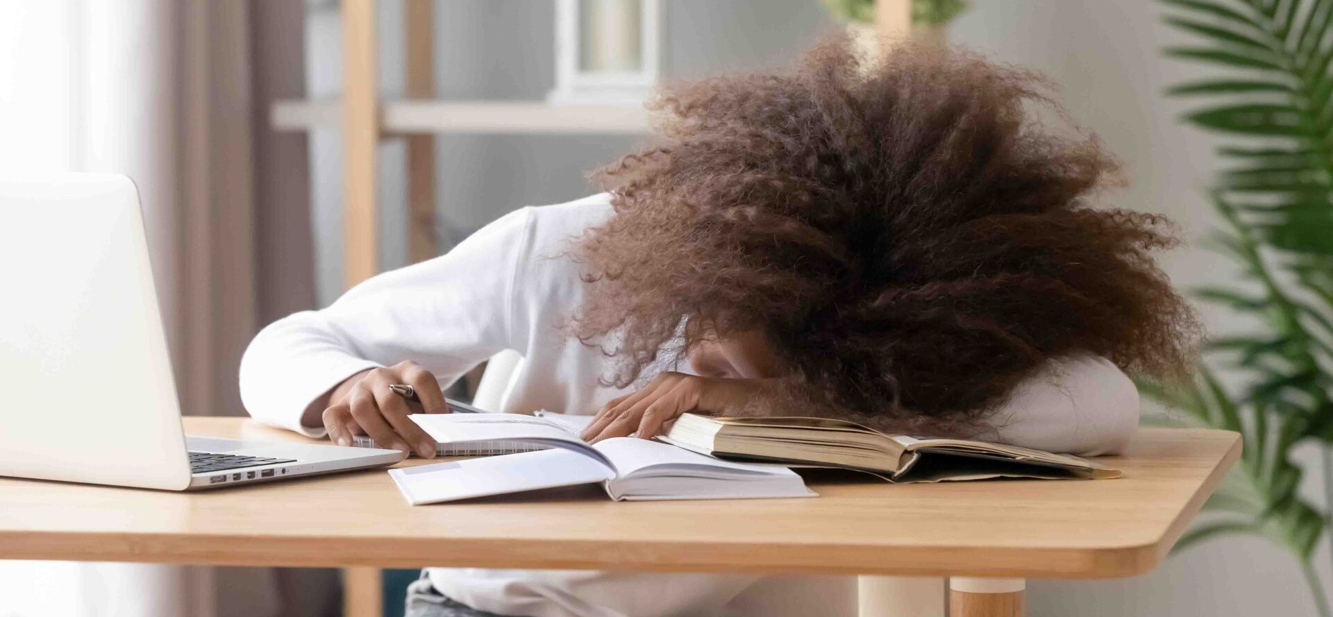 A woman is sleeping at a desk with a laptop and books.