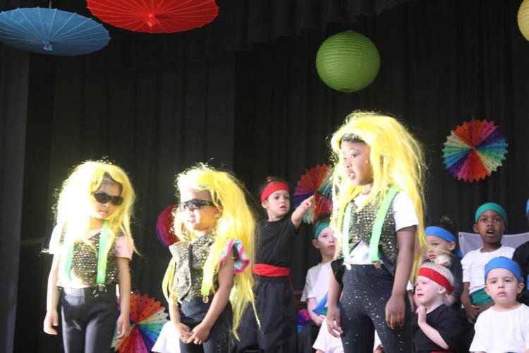 A group of children wearing wigs and sunglasses stand on a stage