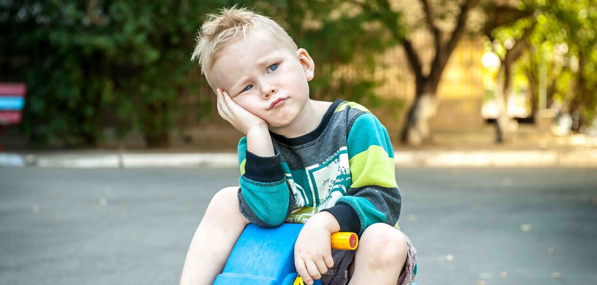 A young boy is sitting on a blue toy car with his hand on his face.