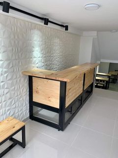 Mostrador largo de madera con base de metal negro en una habitación con azulejos blancos, con un pequeño banco cerca.