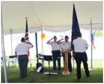 Four men in white shirts salute flags under a tent outdoors.