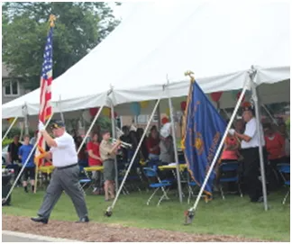 Veterans present flags at outdoor event under a white tent. People, flags, and a scout in the background.
