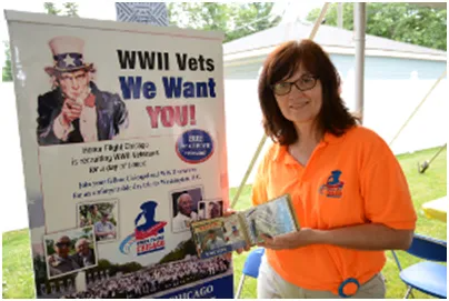 Woman in orange shirt holding items stands by a WWII veterans recruitment banner.