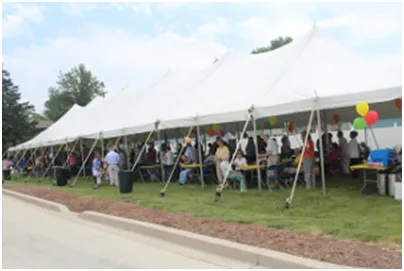 Large white tent with people gathered at an outdoor event, tables and balloons visible.