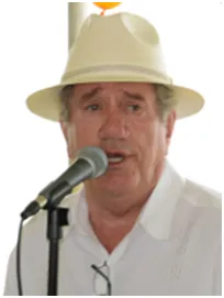 Man speaking into a microphone, wearing a white hat and shirt; outdoors.