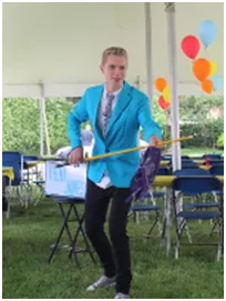 Young man in a blue blazer holding a pole with a purple cloth outdoors with balloons and chairs.