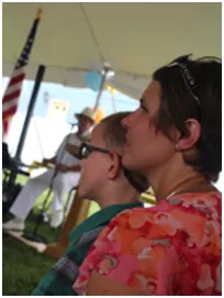 Woman and child watch a musician under a tent, with an American flag in the background.