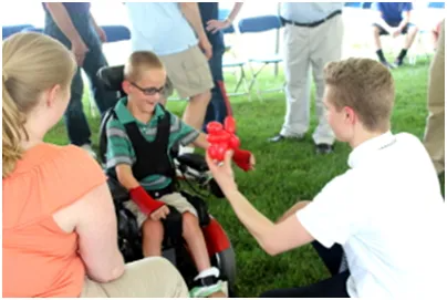 Boy in wheelchair receiving a balloon animal from another person, in an outdoor setting.