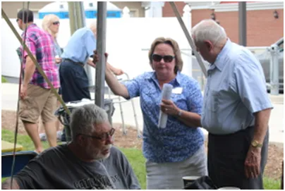 People at outdoor event: woman holding paper, talking with man. Others nearby under canopy.