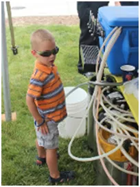 Young child in sunglasses and orange striped shirt looks towards equipment at an outdoor event.