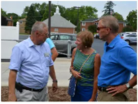 Three people talking outside; a man in blue shirt, a woman in green striped top, and a man in a blue polo shirt.