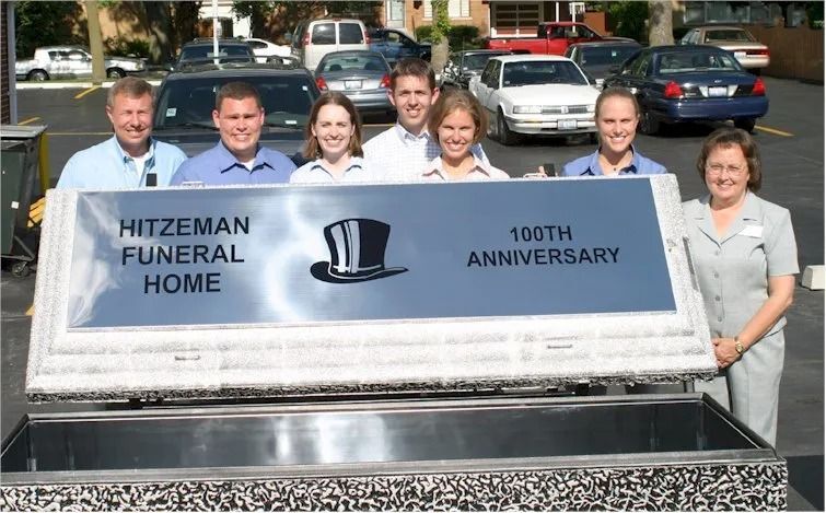 People holding a large plaque for Hitzeman Funeral Home's 100th anniversary, displayed over a casket.