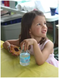 Young girl in pink dress looks upward, holding water bottle and donut at yellow table.