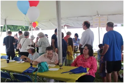 People gather under a tent with balloons. Some are eating at a table covered in yellow.