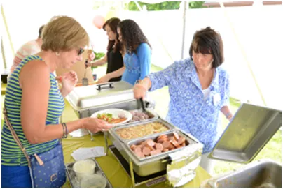 People at a buffet table; woman serving food, other people in background, tent outside.