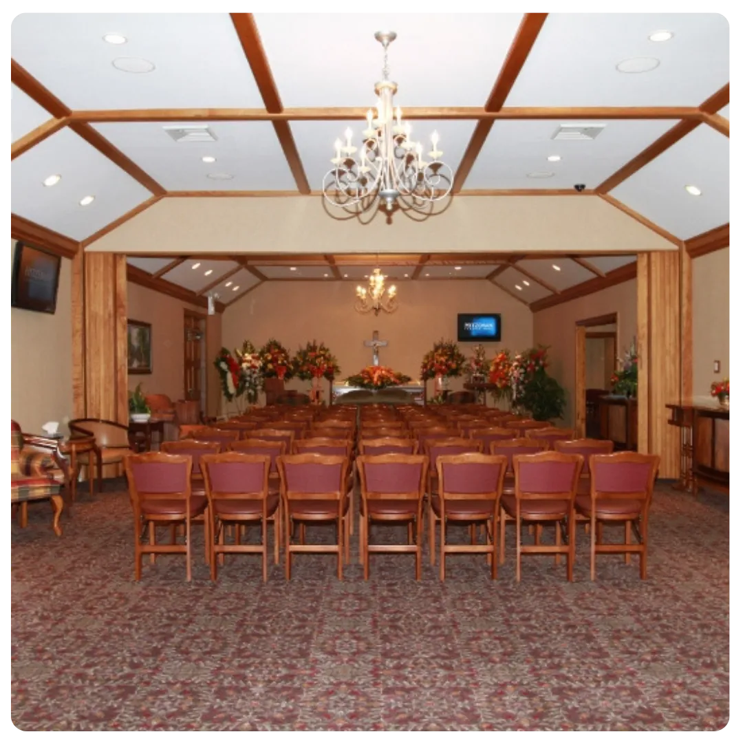 Rows of red chairs face a casket in a funeral home, with floral arrangements and a chandelier.