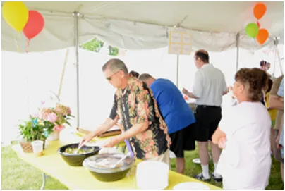 People serving food at a party under a white tent; balloons, flowers, and yellow tablecloth.