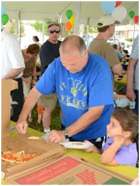 Man in blue shirt serving pizza to a girl at an outdoor gathering. Other people in the background.