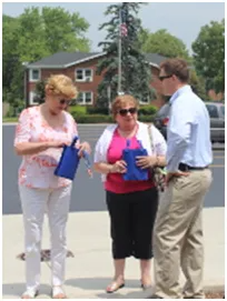 Two women and a man talking outdoors, holding blue bags; building in background.