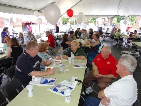 People seated at tables under a tent, socializing and eating at an outdoor event. White and red balloons.