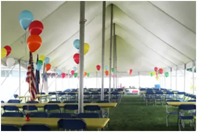 Inside a large white tent, tables and chairs are set up, decorated with balloons.