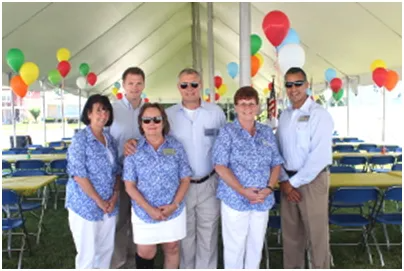 Group of six people smiling under a tent decorated with balloons. They are wearing matching blue floral shirts.