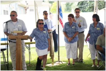 Group of people at podium with flag, some wearing blue floral shirts, outside on grass.